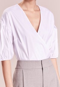 Blusa bianca a V con maniche a sbuffo, realizzata in tessuto morbido. Abbinata a pantaloni grigi a vita alta, sartoriali, con pieghe.
