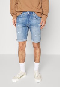 Pantalones cortos de mezclilla azul claro con parches deshilachados y dobladillos enrollados, usados con calcetines blancos y zapatillas beige con bordados florales.