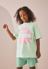 Tricou supradimensionat verde mentă cu imprimeu roz, asortat cu pantaloni scurți cu dungi verzi și albe. Material moale din bumbac, croială casual, mâneci scurte.