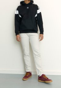 Person som bär en svart och vit Adidas-huvtröja, vita jeans och röda sneakers med gula snören, stående mot en enkel vit vägg.