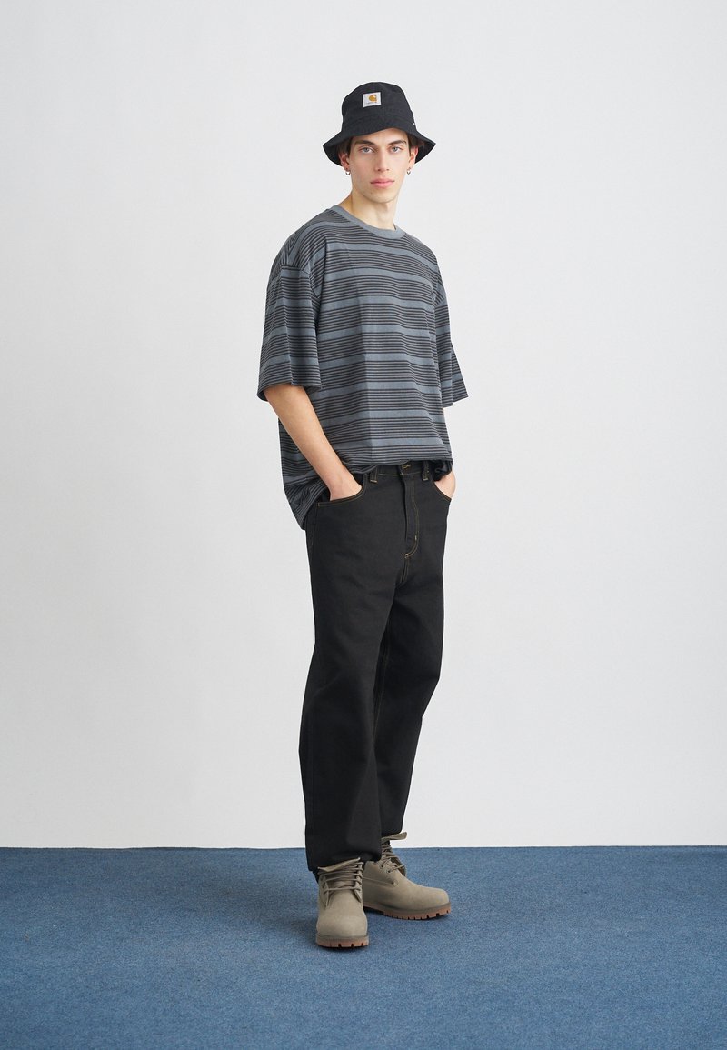 Gestreiftes graues Oversized-T-Shirt, schwarze Jeans und graue Stiefel. Schwarzer Bucket Hat mit Logo-Patch. Lässiges Outfit vor einem blauen Teppichhintergrund.