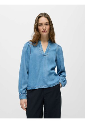 Hosszú hullámos hajú nő világoskék farmer V-nyakú blúzt visel pöttyös hosszú ujjakkal és fekete nadrágot, egyszerű fehér háttér előtt áll.