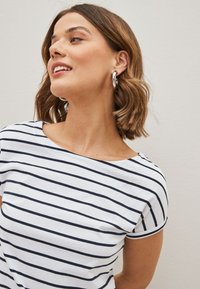 Femme aux cheveux courts et bruns portant des boucles d'oreilles créoles argentées et une chemise blanche à rayures horizontales noires, regardant vers le haut et souriant légèrement.