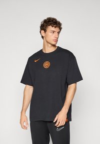 Musta puuvillainen t-paita oranssilla Nike-logolla ja ympyränmuotoisella oranssilla symbolilla rinnassa, jossa on rento malli ja lyhyet hihat.