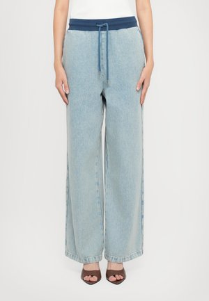 Lyseblå vide jeans med marineblå snøring i livet, brukt sammen med brune åpne hæler mot en hvit bakgrunn.