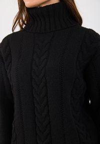 Mustas soonitud sviiter kõrge soonikuga turtleneck'iga, millel on tekstuuriga kaabelmusterd ja vertikaalsed jooned, loodud sooja ja stiili jaoks.