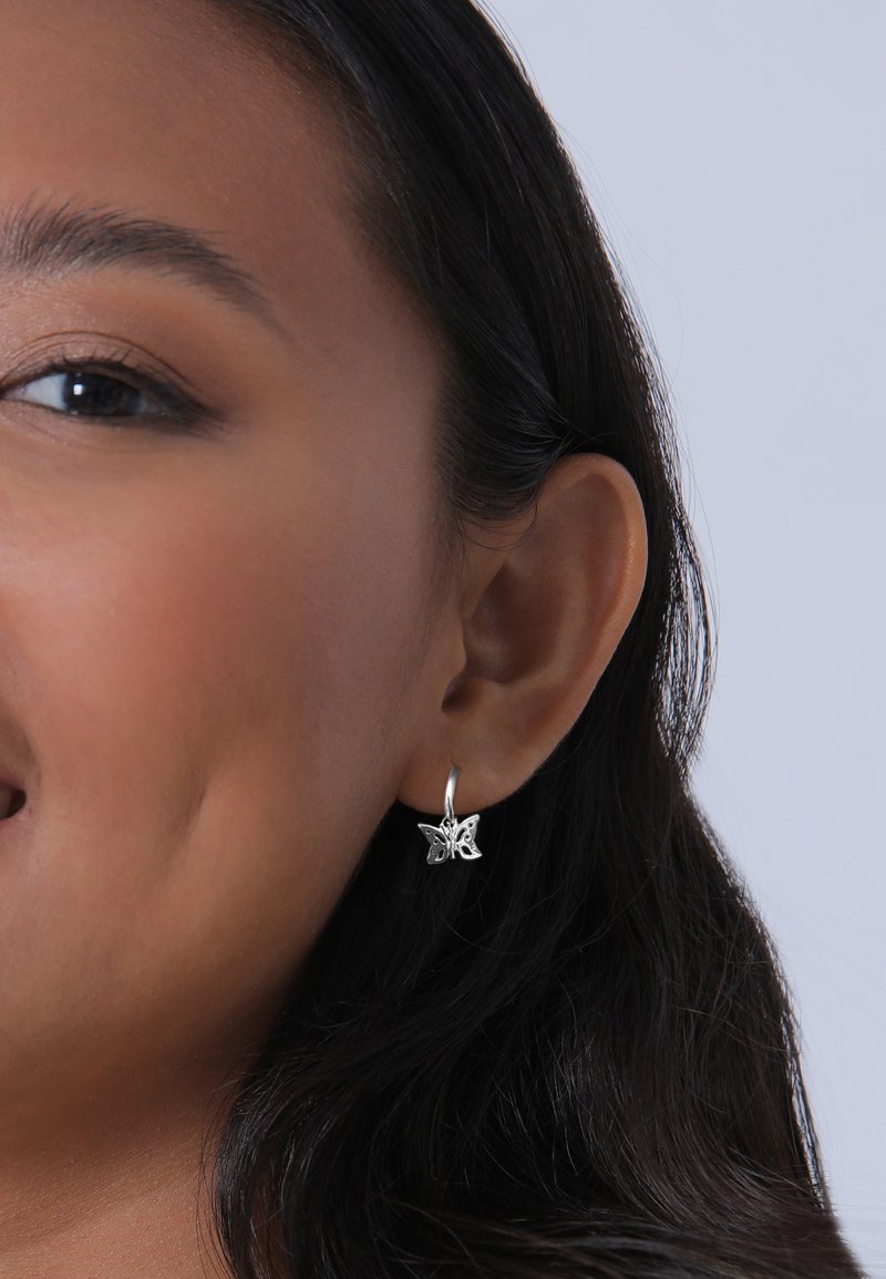 Silverfärgade fjärilsörhängen med detaljerat mönster, hängande från en tunn ring, presenterade i en modells öra mot en ljus bakgrund.