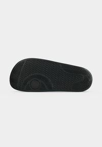 Semelle en caoutchouc noir de tongs avec motifs circulaires, chevrons et lignes texturés pour l'adhérence, et un petit logo circulaire près du talon.