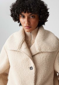 Beige Schaffellmantel mit großem Kragen und einem einzelnen Knopfverschluss. Verfügt über einen gerippten Strickkragen. Weiche Textur und warme Optik.