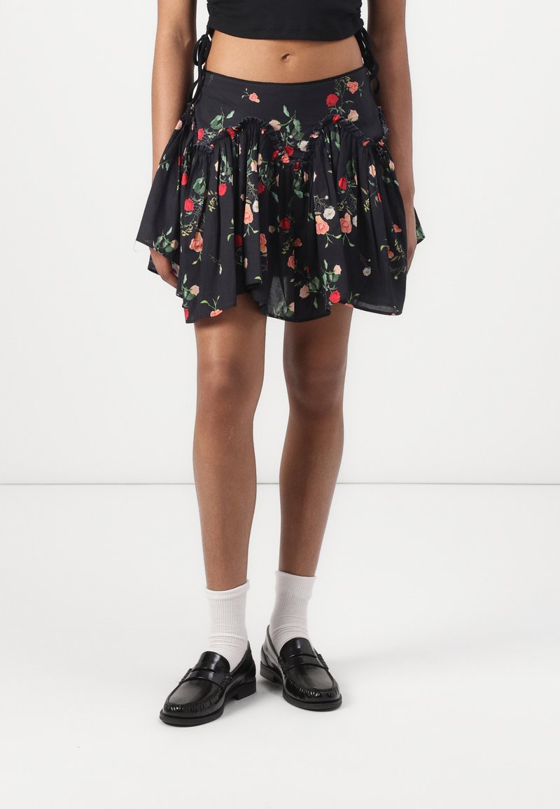 Mini-jupe noire à fleurs avec des détails froncés, portée avec des chaussettes blanches et des mocassins noirs, associée à un top court noir.