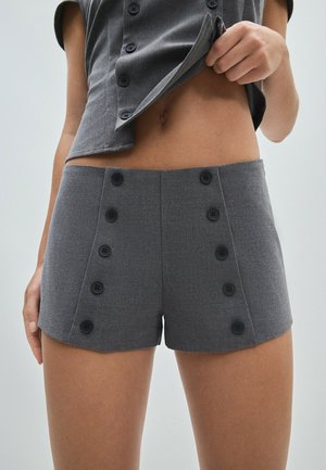 Femme soulevant une chemise grise boutonnée pour montrer un short gris taille haute avec deux rangées verticales de boutons noirs à l'avant.