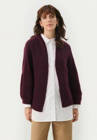 Cardigan lavorato a maglia color bordeaux con una vestibilità morbida, apertura frontale e maniche lunghe. Il tessuto appare morbido e soffice, indossato sopra una camicia bianca.