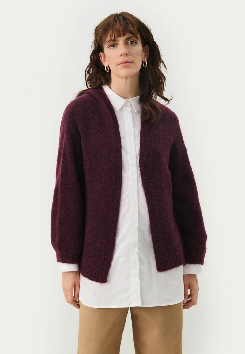 Cardigan lavorato a maglia color bordeaux con una vestibilità morbida, apertura frontale e maniche lunghe. Il tessuto appare morbido e soffice, indossato sopra una camicia bianca.