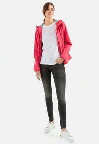 Veste rose déperlant avec capuche, doublure grise, fermeture éclair à l'avant, associée à un t-shirt blanc et un jean foncé, portée avec des baskets blanches.