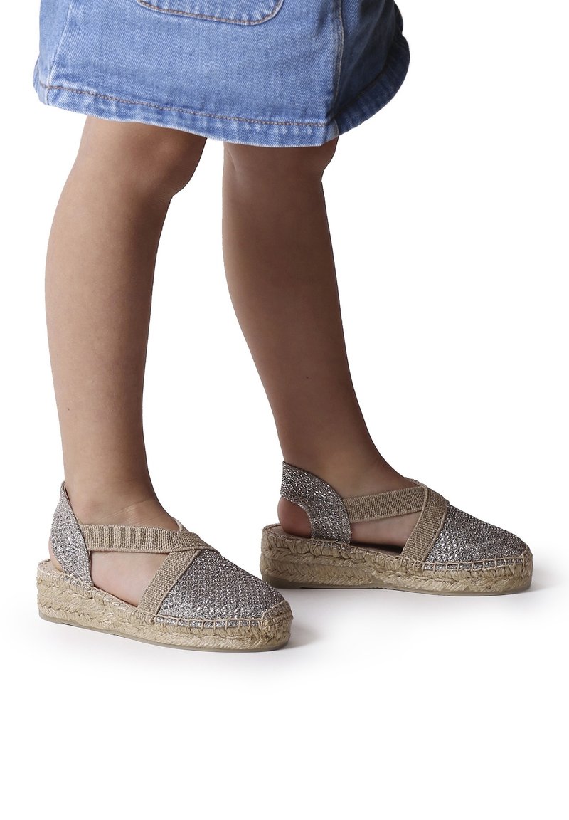 Enfant portant des sandales espadrilles argentées scintillantes avec des brides élastiques beiges et des semelles en jute tressée, associées à une jupe en jean.