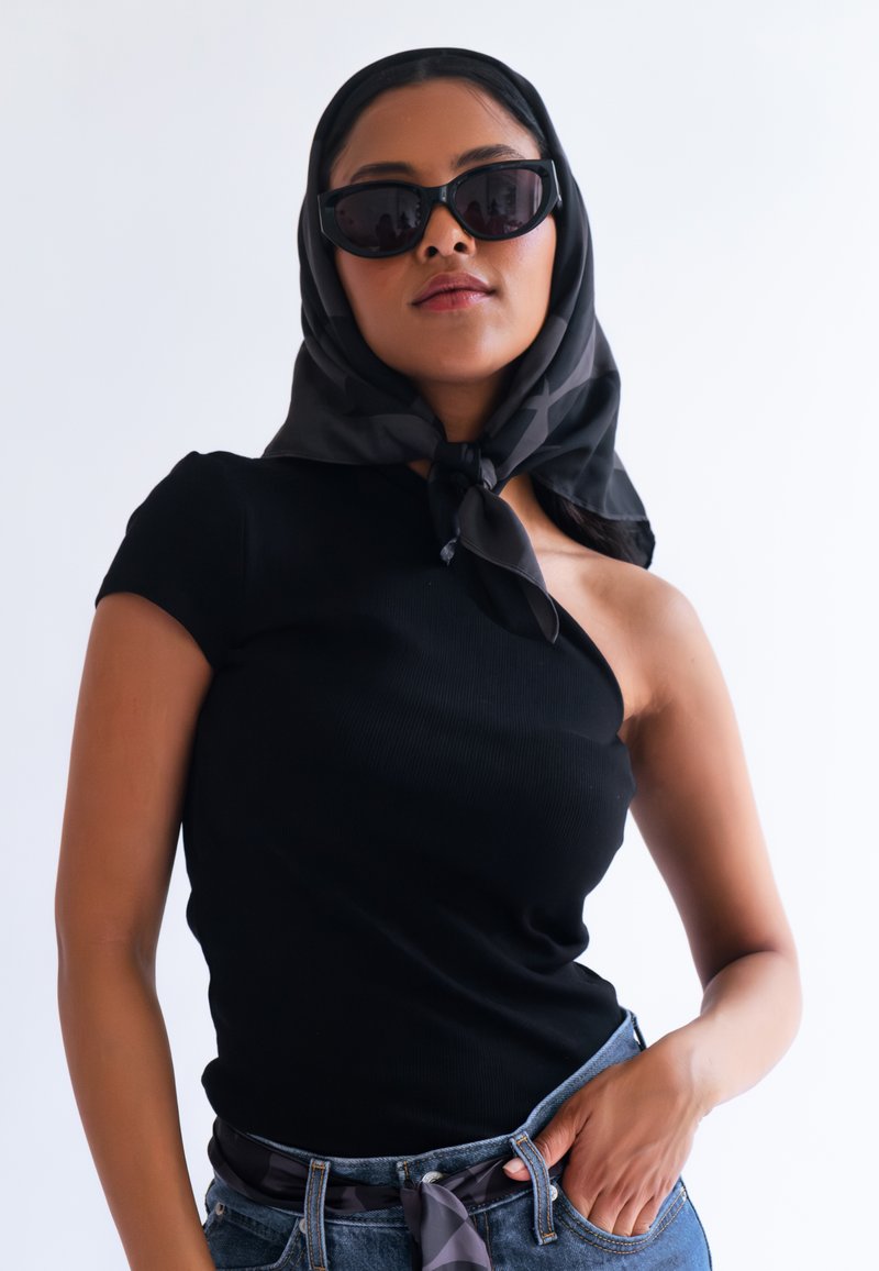 Haut noir à côtes avec une conception à une épaule, associé à un jean en denim et une écharpe noire à motifs nouée au cou. Lunettes de soleil sombres portées.