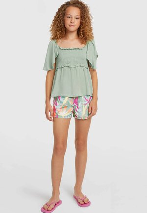 Grünes Oberteil mit Kräuselung und kurzen, flatternden Ärmeln; kombiniert mit bunten Shorts im Blumenmuster. Dazu pinke Flip-Flops.