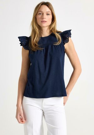 Kvinde med lyst, glat hår iført navyblå ærmeløs top med flæset blonder på skuldrene og hvide bukser, stående foran en ensfarvet baggrund.