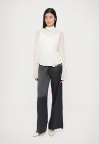 Witte, doorschijnende blouse met ruches aan de halslijn en lange mouwen; gecombineerd met donkergrijze, wijde broekspijpen. Model draagt beige puntige schoenen.