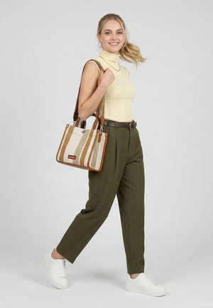 Noor naine kollases varrukateta kaelusega kampsunis, oliivrohelised püksid, valged tossud, triibulise beeži ja helepruuni õlakotiga, naeratades otse vaatajale.