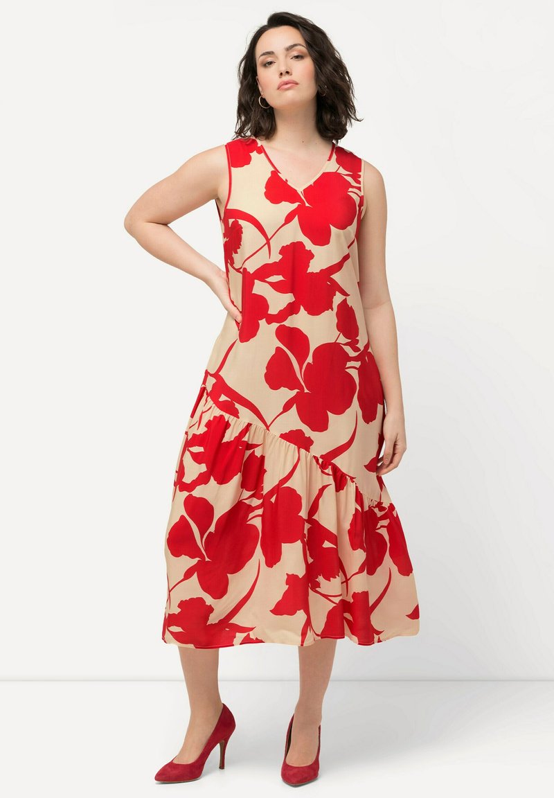 Bezramenné, délky po kolena, bé�žové šaty s velkým červeným květinovým vzorem. Mají výstřih do V a vrstvený volánkový lem. Doplňují je červené lodičky.