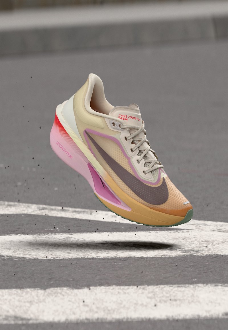 Ανοιχτό μπεζ και ροζ παπούτσι για τρέξιμο Nike Zoom Fly στον αέρα πάνω από άσφαλτο με λευκές οδικές σημάνσεις.