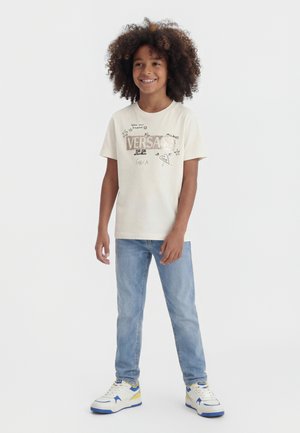 Smilende barn med krøllet hår iført en hvid t-shirt med grafik, lyseblå jeans og hvide sneakers med blå og gule detaljer.