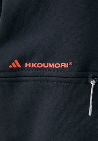 Sort sweatshirt med en lynlåslomme, tekstureret stof og rød branding "H.KOUMORI" med et logo. Kontrastsyninger er synlige.