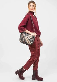 Bolso de hombro con un patrón abstracto en negro, marrón y crema. La modelo lleva un suéter burdeos y pantalones ajustados a juego, con botas negras.