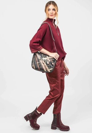Bolso de hombro con un patrón abstracto en negro, marrón y crema. La modelo lleva un suéter burdeos y pantalones ajustados a juego, con botas negras.