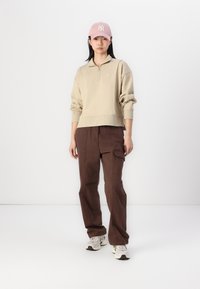 Felpa beige con zip a quarto, pantaloni cargo marroni con tasca laterale e un cappellino rosa con logo. Le sneakers completano l'outfit.