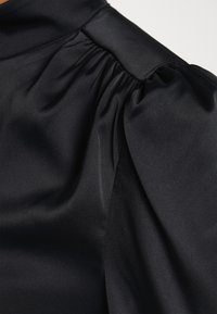 Top de cetim preto com mangas bufantes franzidas, textura suave e um decote alto com costuras limpas nos ombros e no pescoço.