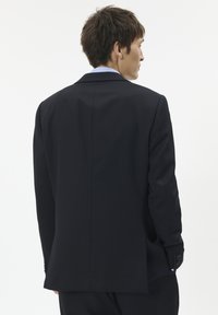 Rövid barna hajú férfi fekete öltönyben és világoskék ingben, háttal állva egy sima fehér háttér előtt.