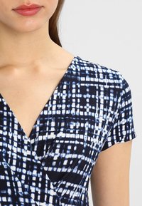 Niebieska i biała sukienka w wzory z dekoltem w kształcie litery V, krótkimi rękawami oraz miękką teksturą materiału z wzorem w kratę.