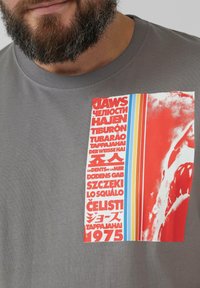 Graues Baumwoll-T-Shirt mit einem auffälligen Grafikdruck, der roten Text und ein Haifischbild zeigt, akzentuiert durch blaue und orange Streifen.