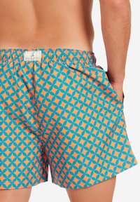 Bunte Badehosen mit einem orange-blauen geometrischen Muster, elastischem Taillenbund und Seitentasche. Der Stoff wirkt leicht und glatt.