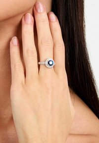 Anello in zaffiro con una pietra centrale blu, di forma rotonda, circondata da un'aureola di piccoli diamanti brillanti. Band in argento con una finitura testurizzata.