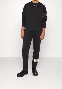 Černé triko s dlouhým rukávem s bílým designem tří pruhů na rukávech, doplněné černými zúženými kalhotami s bílými pruhy u kotníků. Černé robustní boty.