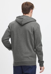 Man in een grijze hoodie en marineblauwe broek, staande met de rug naar de camera tegen een effen witte achtergrond.