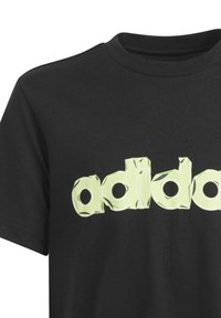 Mustapaita, joka on valmistettu puuvillasta, ja jossa on suuri, teksturoitu keltavihreä "adidas"-logo rinnassa lohkomaista, tyyliteltyä muotoilua.