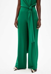 Groene jumpsuit met brede pijpen van soepel materiaal, met een knoopdetail aan de taille. Bevat puntige schoenen met een zwart-wit koeienprint. 