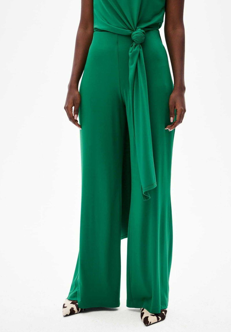 Groene jumpsuit met brede pijpen van soepel materiaal, met een knoopdetail aan de taille. Bevat puntige schoenen met een zwart-wit koeienprint. 