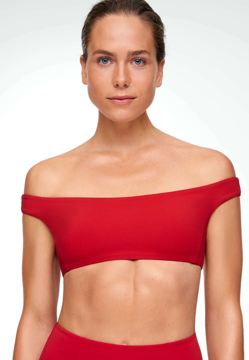 Femme aux yeux bleus et cheveux tirés en arrière, portant un crop top rouge à épaules dénudées sur fond blanc.