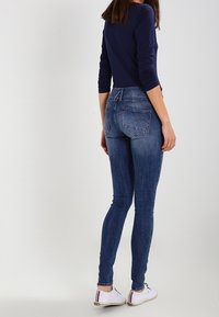 Jeans ajustados en denim azul con un acabado desvanecido. Presentan bolsillos traseros y detalles de costura sutiles. Combinados con zapatos blancos.
