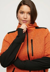Giacca a zip arancione con maniche nere, dotata di colletto alto, tasca sul petto con zip e un design aderente.