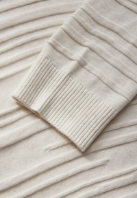 Krämmig stickad tyg med texturerade, upphöjda ränder och ribbade ärmar. Materialet verkar mjukt och lätt.