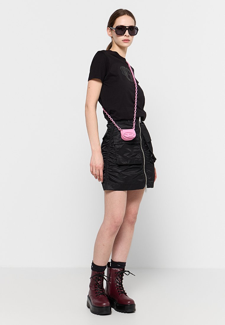 T-shirt nero, gonna nera con zip, borsa a tracolla rosa con catena. Il modello indossa occhiali da sole scuri e stivaletti borgogna con lacci spessi.