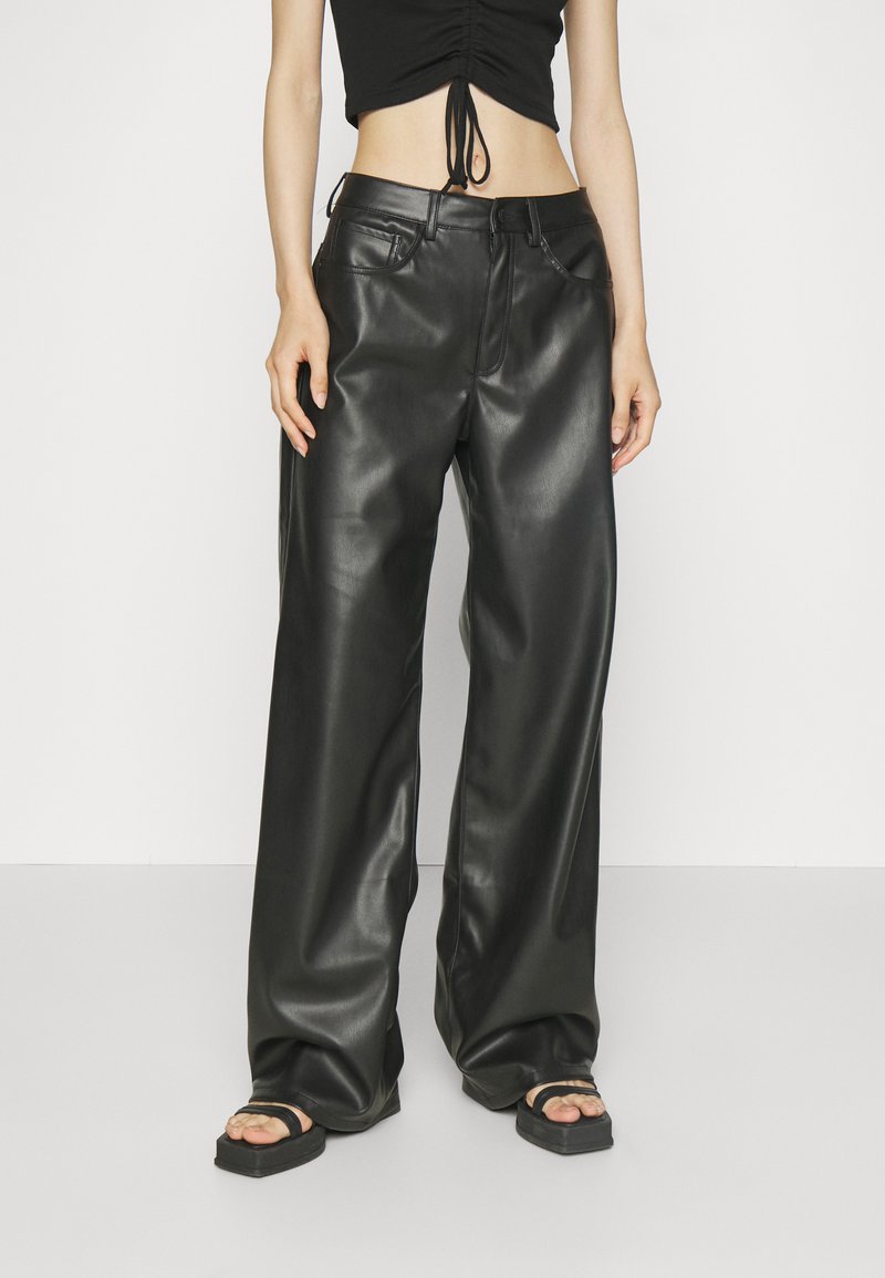 Hoge taille zwarte leren wijde broek met een gladde textuur, knoopsluiting en riemlussen. Draagt met een cropped top en sandalen.