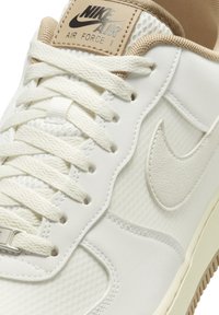 Bílé tenisky Nike Air Force 1 s texturovanou kůží a síťovinou, se béžovými akcenty, plochými bavlněnými tkaničkami a logem na jazyku.