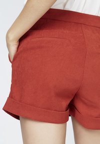 Shorts en daim rouge avec un ourlet roulé et une poche arrière, présentant une texture lisse et une coupe ajustée.